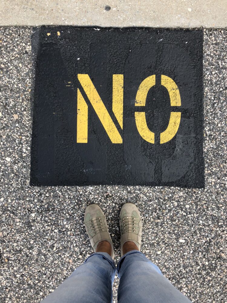 Ein Schild mit dem Inhalt NO (deutsch: Nein) auf schwarzem Grund in gelber Schrift, davor eine Person deren Schuhe man sieht