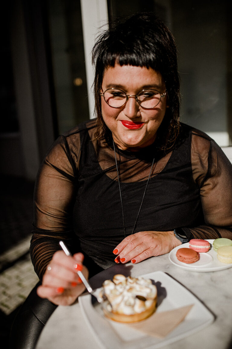 Sabine sitzt in einem Kaffee und ist lächelnd ein Stück Kuchen
