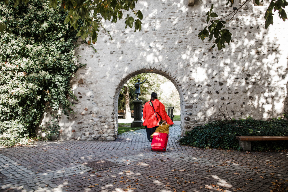 Eine Frau geht durch ein mittelalterliches Tor und zieht einen schweren Koffer hinter sich her. Sinnbild, um das Helfersyndrom zu überwinden.